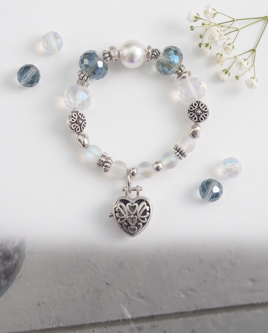 A beaded bracelet with a heart-shaped silver pendant, featuring faceted blue beads, iridescent white beads, clear glass beads, and silver spacer details, styled on a white surface with small white flowers.